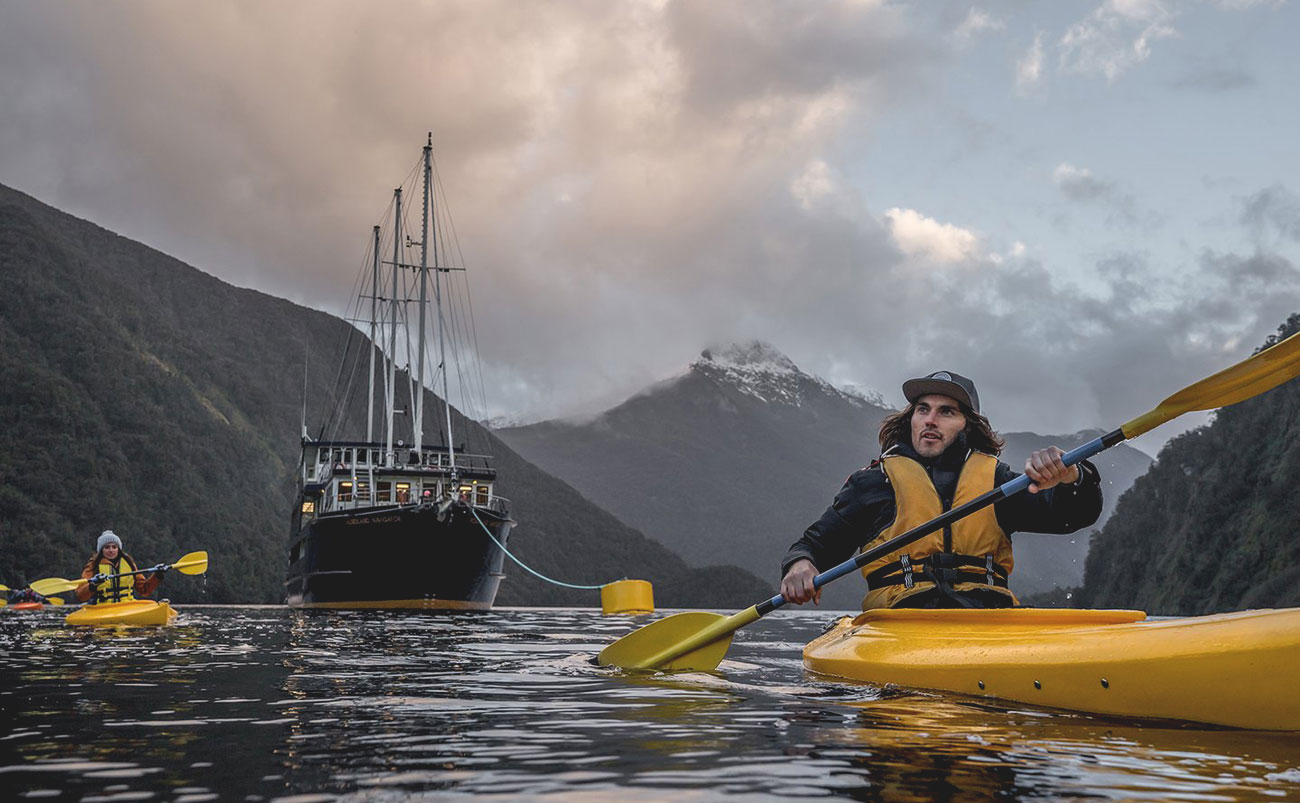 Kayakers paddling through the calm waters of Doubtful Sound, Fiordland, with steep forested cliffs rising from the water, New Zealand