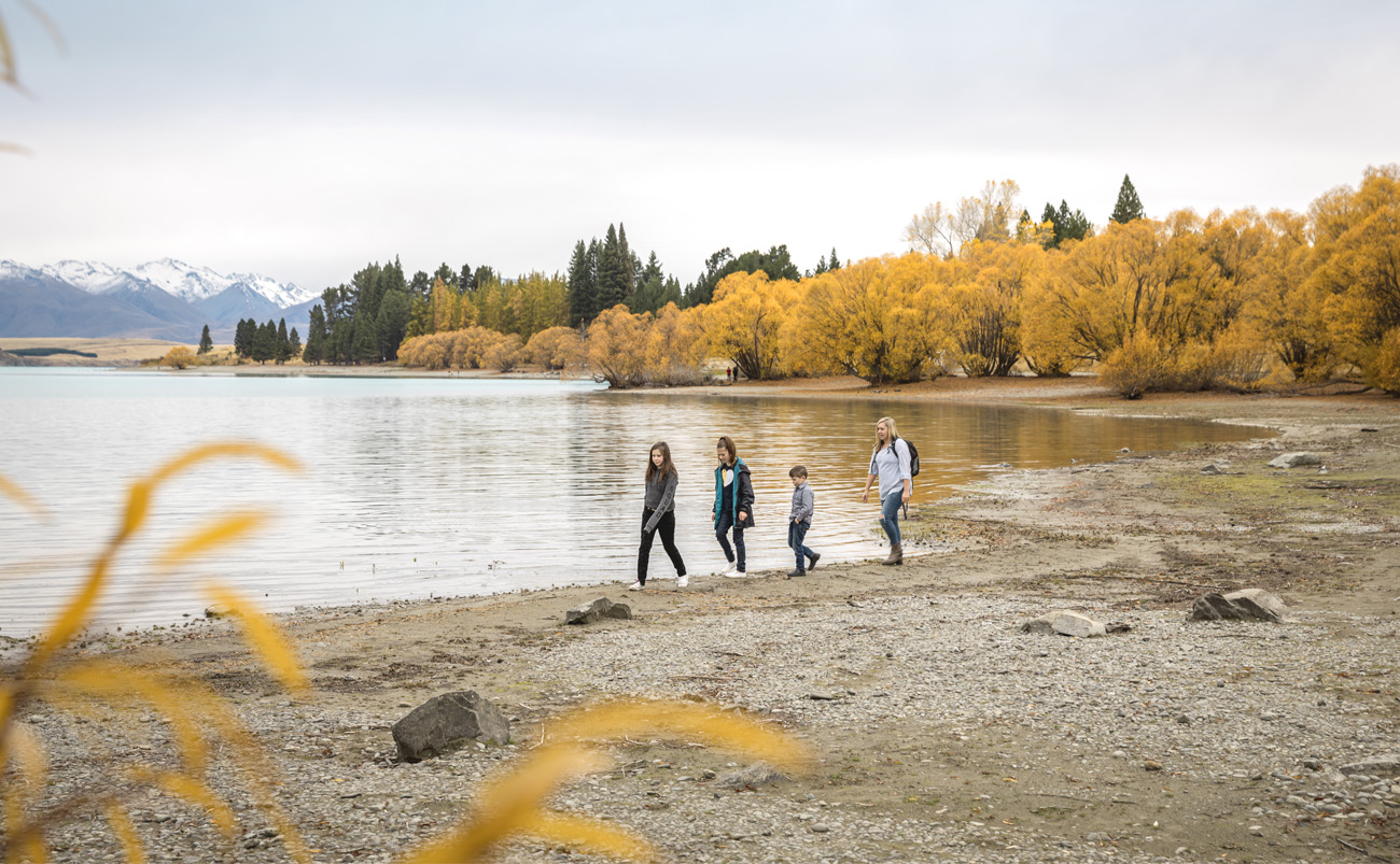 Family walking beside Lake Tekapo in autumn with turquoise water, golden hills and snow-capped mountains in the distance